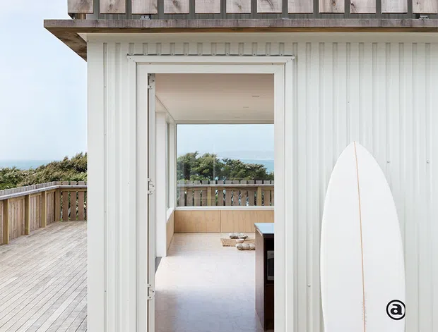 Exterior view of a coastal-style building clad in white vertical corrugated metal, opening onto a timber deck. A weathered timber privacy screen or upper-level balustrade sits above the roofline, while a surfboard leans against the wall beside the doorway. Through the open door, a bright interior and ocean-facing outlook are visible.