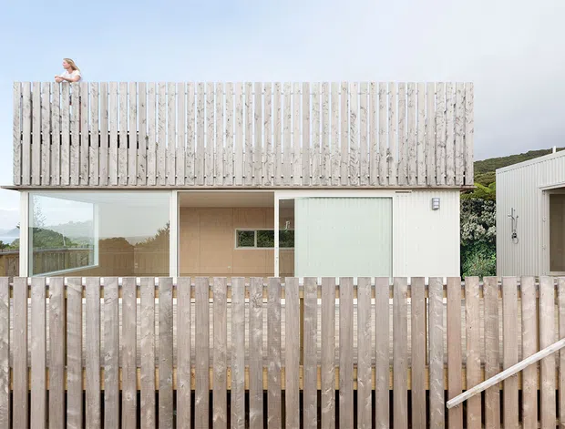 Exterior view of a contemporary two-storey coastal home with extensive vertical timber privacy screening and fencing. Large glazed openings reveal a simple interior, while a separate light-coloured auxiliary structure sits to the right. The elevated timber enclosure partially obscures the lower level, and a glimpse of ocean and hillside is visible beyond.
