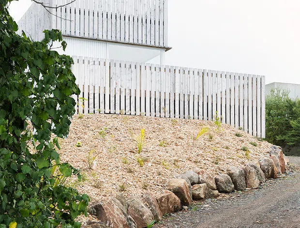 Exterior view of a contemporary multi-level house set into a landscaped, rocky site. The building features pale vertical timber slat screening and cladding, a simple flat-roofed cubic form, and a partially concealed upper volume. Gravel paths, stone retaining edges, and established trees surround the structure.