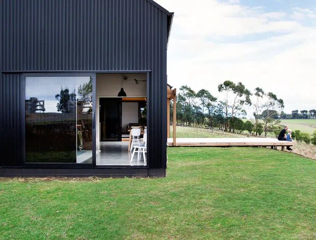 Exterior view of a compact contemporary gabled building clad in dark vertical corrugated metal, with large glazed openings revealing a simple interior. The building sits in an expansive grassy rural landscape, with a timber deck or walkway extending along the side and open farmland beyond.