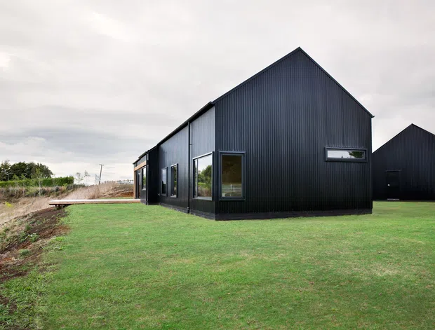 Exterior view of a contemporary single-storey building with steep gable roofs and dark vertically ribbed metal cladding. The structure has simple rectangular window openings with dark frames and sits on a broad grassed site, with a smaller matching gabled outbuilding behind it. The overcast setting and restrained black-and-grey palette create a clean, understated architectural character.
