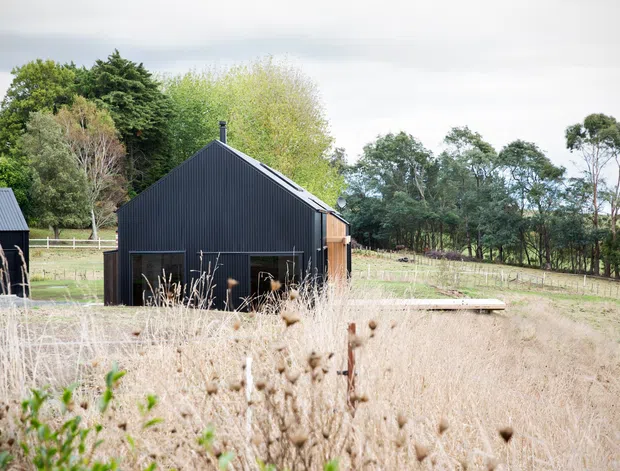 A small dark-clad gable-roofed building sits in a rural pastoral landscape, surrounded by dry grasses, mature trees, fencing and paddocks. The simple structure has vertically ribbed black metal cladding, a steep pitched roof, dark-framed openings and a timber-clad side section. A second matching outbuilding is partially visible at left.