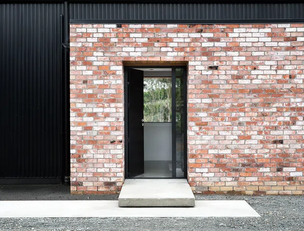 Front elevation of a simple industrial-style building with a pale corrugated metal pitched roof, black vertically ribbed metal cladding, and a prominent weathered red-and-white brick wall. A dark glazed entry door sits centrally within the brick facade, accessed by a small concrete step and path, with a gravel forecourt.