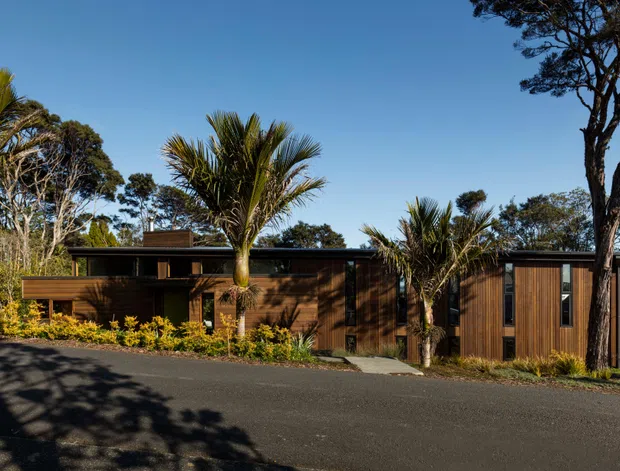 Single-storey contemporary timber-clad house viewed from the street, integrated into a leafy landscaped setting. The low horizontal composition features dark wood walls, broad rooflines, vertical timber screening, extensive glazing, palms and native-looking trees, with a paved driveway or road in the foreground.