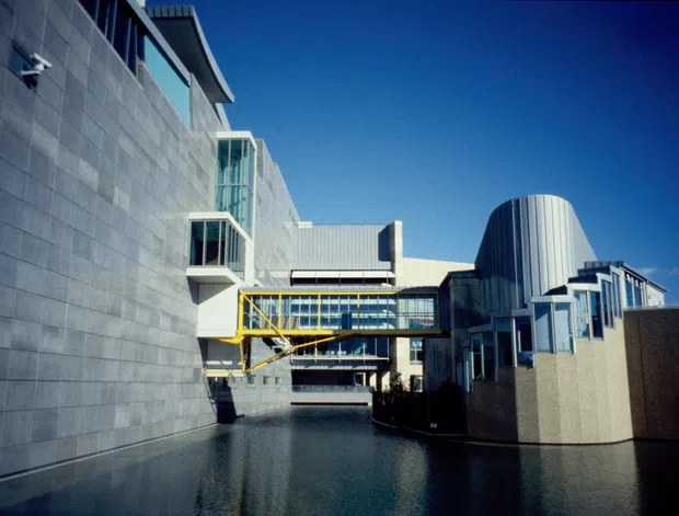 Exterior view of a large modern institutional or commercial building complex with strong geometric forms. The composition shows a broad grey tiled wall, glazed elevated link bridges with yellow structural framing, projecting balconies, vertical window bays, and a prominent curved ribbed metal-clad volume. A shallow reflecting pool occupies the foreground under a clear blue sky.