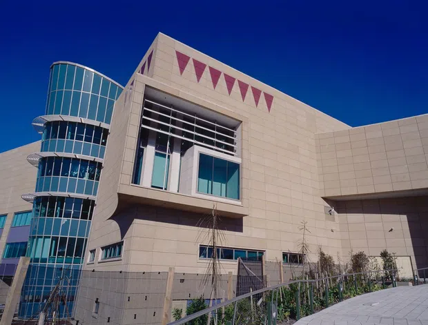 Exterior view of a large contemporary institutional or commercial building under a deep blue sky. The facade combines pale stone or concrete-like cladding, extensive blue-tinted curtain-wall glazing, a rounded glazed corner tower, projecting rectangular volumes, horizontal sun-shading elements, and a large recessed entrance or covered forecourt. Landscaping and paving occupy the foreground.