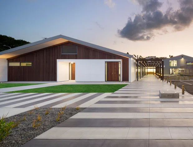 Wide dusk exterior view of a contemporary low-rise building complex. The foreground features a broad pedestrian plaza with alternating light and dark linear paving, gravel planting beds, bollards, and a covered timber-framed walkway leading toward additional buildings. The main single-storey gabled building has a white rendered or panelled facade, extensive vertical dark timber cladding, a white garage-style door, and timber entry doors. Additional modern buildings and parking are visible to the right.