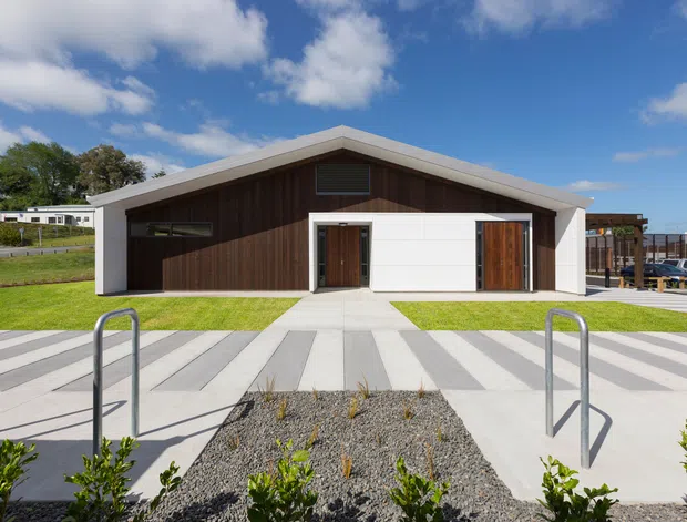 A low, single-storey contemporary building with a strong asymmetrical gable roof and a contrasting facade of dark vertical timber cladding and crisp white panels. Timber entrance doors, a narrow horizontal window, and high-level ventilation openings face a broad patterned concrete forecourt. The foreground includes pool-style handrails and gravel planting beds, while a deck or covered structure and parked vehicles are visible to the side. The scene is brightly lit under a blue sky.