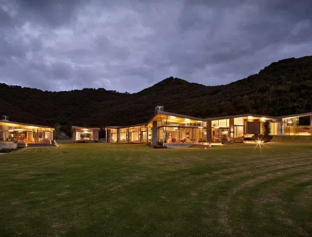 A sprawling single-storey modern residence set against wooded hills, photographed at dusk from a broad manicured lawn. The house has low-pitched rooflines, extensive floor-to-ceiling glazing, warm interior lighting, and multiple connected wings arranged around outdoor living areas.