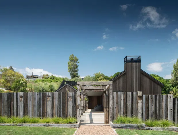 Front exterior view of a low-rise residential building behind a tall, weathered timber privacy fence. The home has a strongly pitched gable roof, dark vertical timber cladding, and a prominent chimney or screened rooftop element. A central timber-framed pedestrian entry opens through the fence to a paved path, with landscaped grasses and shrubs along the frontage.