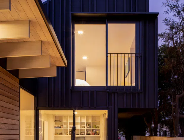 Twilight exterior view of a modern multi-level residence. The building combines dark vertical metal cladding with warm timber-lined soffits and timber wall cladding. Floor-to-ceiling glazing reveals a warmly lit home office or study with built-in shelving and a lounge chair, while an upper-level glazed opening includes a narrow balcony with a dark metal balustrade. A timber deck and walkway connect the foreground to the house, with landscaped planting and a boundary fence at the side.