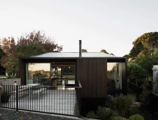 Exterior view of a compact modern single-storey house or extension set within established landscaping. The building has dark vertical timber cladding, large glazed openings, a simple low-pitched metal roof with a prominent flue, and a raised deck or terrace enclosed by black metal balustrades. The glazing reveals a contemporary kitchen or living area inside, while native-style planting surrounds the structure.