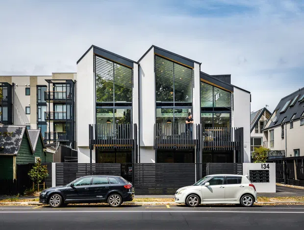 Street-facing view of a contemporary multi-unit residential building with three tall gabled facade bays, extensive upper-level glazing, horizontal blinds, dark vertical balcony screens, and black slatted fencing. Adjacent homes and parked cars line the urban residential street.