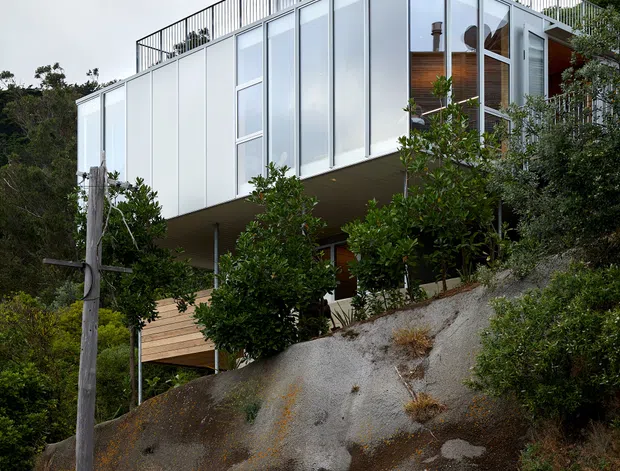 Exterior view of a contemporary multi-level house perched on a steep rocky, vegetated site. The building features extensive floor-to-ceiling glazing, pale metal panel cladding, a projecting upper volume, timber decking, and a rooftop terrace enclosed by a simple metal balustrade. A chimney rises above the roof, and the structure appears elevated over the sloping terrain.