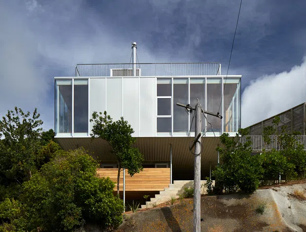 Exterior view of a contemporary elevated dwelling on a sloping, landscaped site. The building has a box-like upper volume with extensive floor-to-ceiling glazing, translucent or opaque light facade panels, a projecting soffit, rooftop metal railing, and a visible flue. Timber screening and steps lead up from the rocky planted foreground, with a neighbouring deck structure at right.