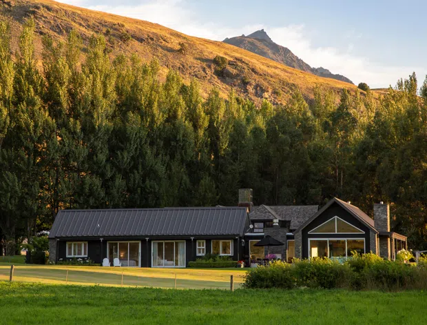 A low, dark-clad rural house set at the edge of a lawn, surrounded by dense trees and backed by a steep golden hillside with a rugged mountain peak. The home has a simple pitched-gable form, large glazed openings, and a contemporary lodge-like appearance.