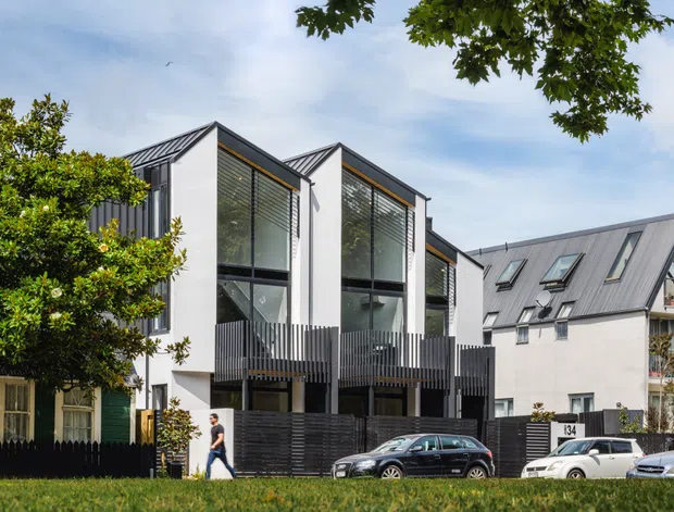 Exterior view of a contemporary multi-storey residential development with sharply pitched gabled roof forms, white rendered walls, dark vertical cladding, large glazed openings with external blinds, and projecting dark timber or metal balconies. A neighbouring pitched-roof building and mature street trees are visible, with parked cars in the foreground.