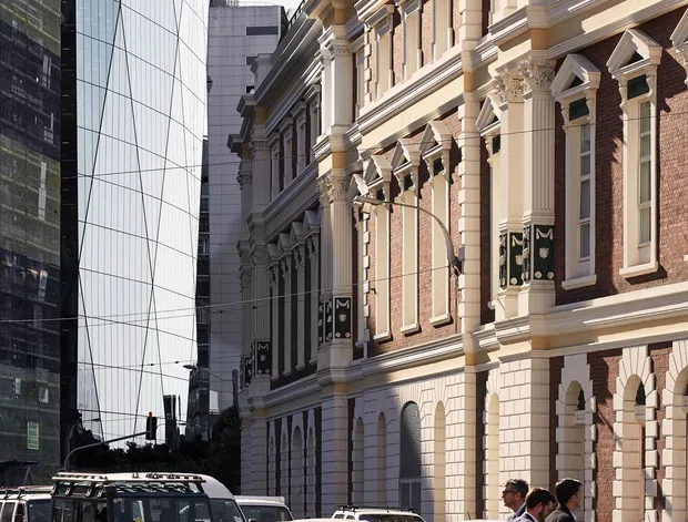 Urban streetscape showing a grand historic brick building with elaborate classical detailing, pilasters, decorative cornices, arched openings, and cream stone trim beside a contrasting contemporary glass office building. Vehicles and pedestrians occupy the foreground street.