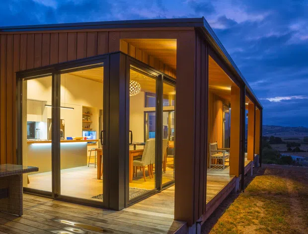Twilight exterior view of a modern single-storey home with extensive floor-to-ceiling glazing, warm timber cladding, and a covered timber deck. The illuminated interior reveals an open-plan kitchen and dining area, while the home sits on an elevated or sloping site overlooking a broad landscape.