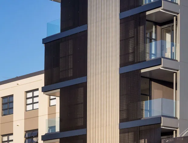 Exterior architectural photograph of a contemporary multi-storey apartment or commercial building. The facade combines dark screening panels, a tall vertically ribbed light cladding element, projecting balconies with clear glass balustrades, dark horizontal bands, and large glazed openings. A neighbouring light-coloured building is visible in the background under a clear blue sky.