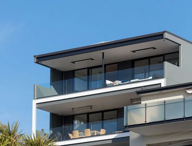 Exterior view of a contemporary multi-storey residential building with stacked balconies, broad roof overhangs, dark-framed glazing, clear glass balustrades, and a light grey facade. Outdoor furniture is visible on the balconies, with neighboring roof forms and planting in the foreground under a clear blue sky.