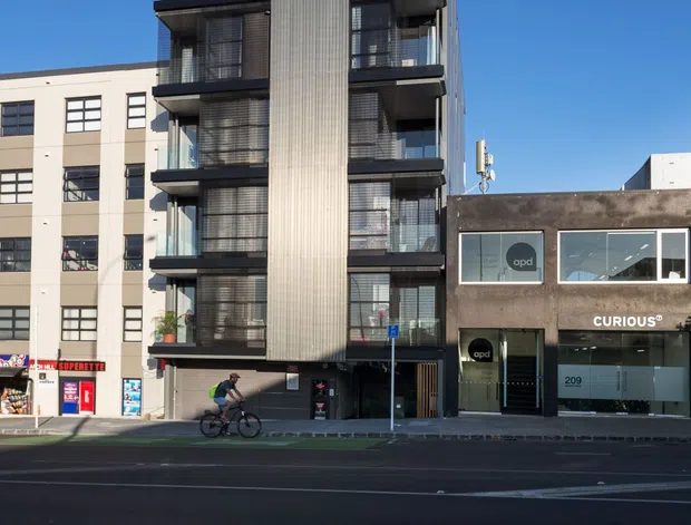 Street-level view of a contemporary multi-storey apartment building in an urban commercial streetscape. The narrow building has several stacked balconies with dark metal balustrades, extensive glazing, and a prominent vertically ribbed light-coloured facade panel. Adjacent low-rise commercial buildings and street-front retail are visible.