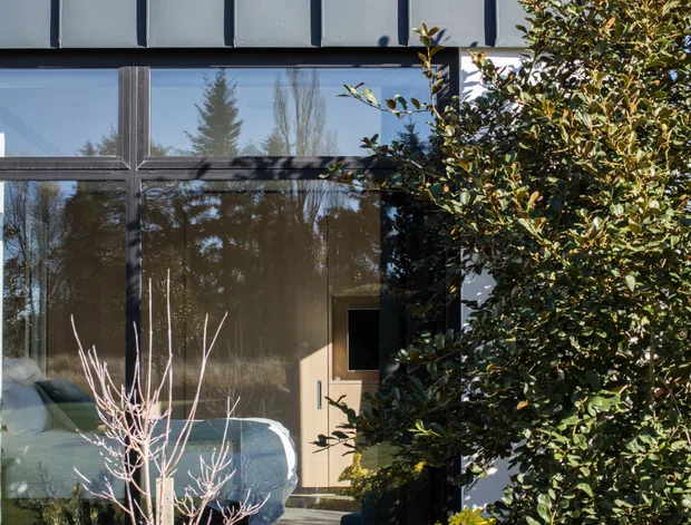 Exterior view of a contemporary multi-level facade with dark framed floor-to-ceiling glazing, a light grey vertically ribbed metal-clad upper wall, and a recessed timber entry door. Dense landscaped planting partially screens the building, while the glazing reflects surrounding trees and reveals a softly furnished interior.