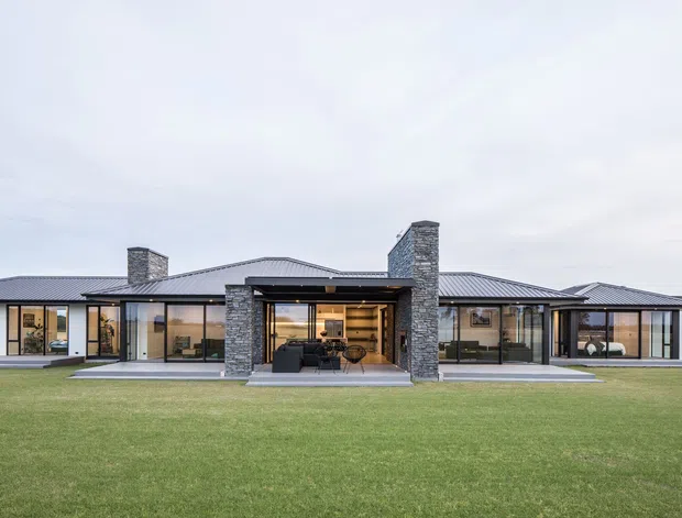 A wide exterior view of a low, single-storey contemporary residence set in an expansive lawn. The home has a long horizontal composition, dark standing-seam metal hipped roof forms, extensive floor-to-ceiling glazing, and several prominent rough grey stone chimney or masonry volumes. A recessed central outdoor living area with furniture is framed by stone piers, while warm interior lighting is visible through the glazing.
