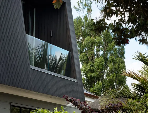 Exterior view of a contemporary multi-level house, dominated by a dark vertically ribbed cladding facade with a steeply angled projecting upper-storey form. A glass-balustraded balcony is partially visible, with large windows at ground level and dense landscaped planting in the foreground.