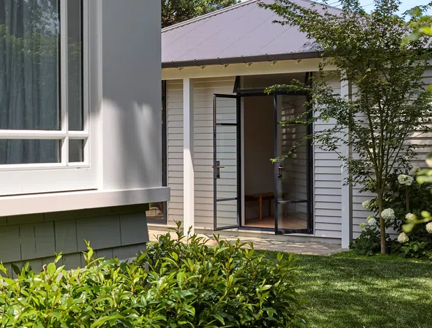 Exterior view of a light-coloured residential building with horizontal weatherboard cladding, a muted metal hip roof, large window, and glazed double doors opening onto a paved garden path. Trimmed hedges, lawn, and mature planting frame the landscaped courtyard-like approach.