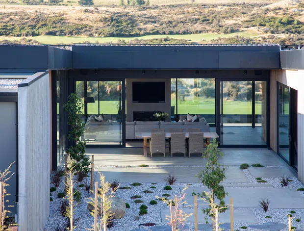 Modern single-storey residence with dark metal cladding, extensive floor-to-ceiling glazing, and a covered outdoor entertaining area opening onto a landscaped gravel courtyard. The visible interior includes a dining table and living area, while the surrounding dry, native-style planting creates a low-maintenance landscape setting.