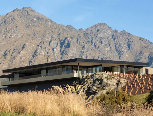 A low, contemporary flat-roofed building set against a dramatic rugged mountain range. The structure has extensive dark-framed glazing, projecting horizontal roof planes, dark exterior cladding, and a terraced or stepped retaining wall on the right. Dry grasses and natural rocky terrain surround the building.