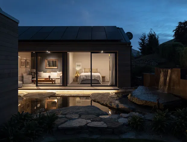 Twilight exterior view of a compact modern dwelling opening onto a landscaped courtyard with a reflective pond and natural stone paving. Dark horizontal cladding and a low pitched roof frame expansive floor-to-ceiling glazing, through which a softly lit bedroom and adjacent living area are visible. The setting has a tranquil, integrated indoor-outdoor character with planted garden beds, stonework, timber screening and warm interior lighting.