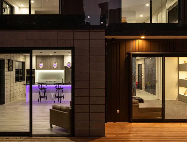 Nighttime exterior view of a contemporary multi-level dwelling with extensive floor-to-ceiling glazing and dark framed sliding doors. Warm timber decking and vertical timber cladding contrast with dark masonry panels. Through the glazing, a softly lit interior with a bar or kitchen counter, stools, pendant lights, built-in display shelving and a seating area is visible, with purple accent lighting.