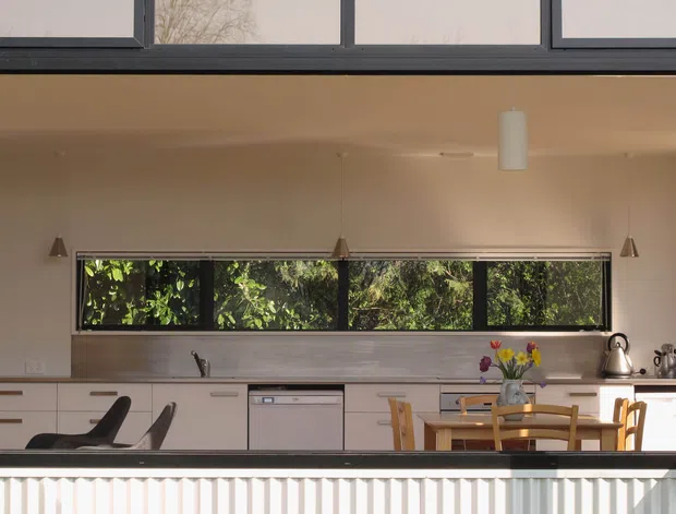 Exterior view through a large open framed window into a bright contemporary kitchen and dining area. The room features a long run of pale flat-panel cabinetry, dark-framed horizontal glazing with garden views, timber dining furniture, pendant lights, and stainless-steel appliances. The building exterior has light corrugated cladding and dark metal window framing.