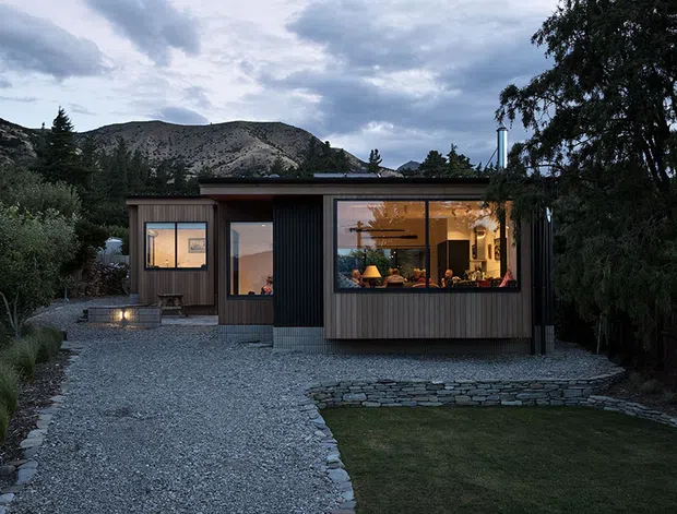 A compact single-storey contemporary home viewed from the landscaped approach at dusk. The dwelling has a low flat roof, dark vertical metal cladding, warm timber-toned wall sections, and large illuminated windows revealing a warmly furnished interior. A gravel driveway and path lead to the house, with stone edging, a small lawn, mature trees, and rugged mountain terrain behind.