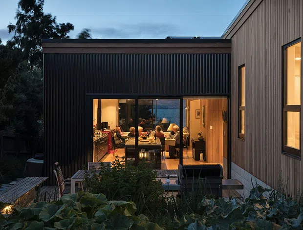 Twilight exterior view of a contemporary multi-volume home, with dark vertically ribbed cladding on the left wing and lighter vertical timber-like cladding on the right. Full-height glazed doors reveal a warmly lit dining and living area inside, while a planted terrace and garden occupy the foreground.