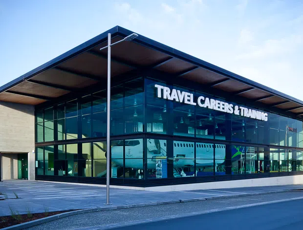 Contemporary two-storey commercial training facility with a broad glazed curtain wall, dark metal-framed flat roof canopy, and contrasting light-coloured solid wall panels. The building signage identifies it as a travel careers and training centre. A paved forecourt and street frontage are visible.