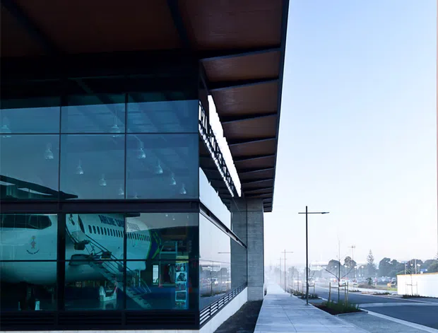 Street-level exterior view of a contemporary public or commercial building with extensive floor-to-ceiling glazing, a deep projecting roof canopy with dark metal framing and timber-toned soffit panels, and a paved pedestrian forecourt. The glass reveals an aircraft display or aviation-themed interior. The building sits along a broad urban streetscape with simple modern street lighting and roadway infrastructure.