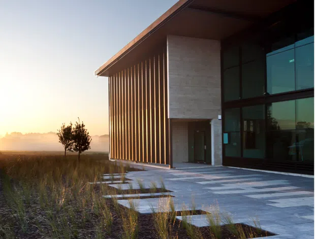 Exterior architectural photograph of a contemporary low-rise building at sunset. The building features a broad projecting roof, extensive dark-framed glazing, a pale concrete wall volume, and a vertical timber or wood-toned sun-screen facade. A paved terrace or entry path of large rectangular slabs crosses a landscaped bed of ornamental grasses, with open rural or natural surroundings beyond.