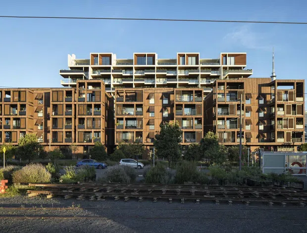 Exterior view of a contemporary multi-storey apartment building with a warm brown timber-toned facade, extensive balconies, large windows, and stepped rooftop structures. The building sits in an urban setting beside railway tracks, with landscaping and parked cars in the foreground.
