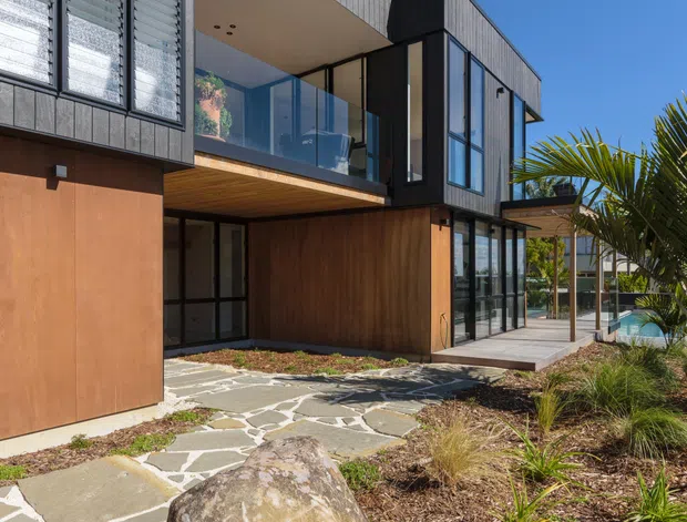 Exterior view of a contemporary multi-level home with dark grey fibre-cement or panel cladding, warm timber-toned wall panels, extensive glazing, louvred windows, projecting balconies, and a covered outdoor terrace. A stone-paved path, native-style landscaping, grasses, and a large boulder occupy the foreground, with a glimpse of a pool or water feature at the side.