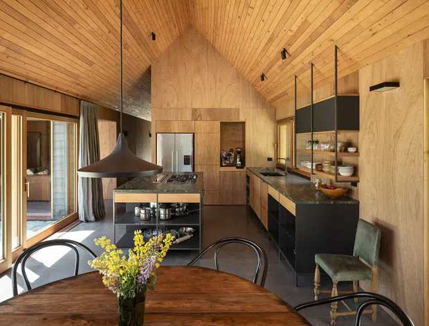 Warm, timber-lined contemporary kitchen and dining space with a dramatic pitched timber ceiling. The kitchen features dark cabinetry, stone benchtops, a central island with open shelving and cooktop, integrated stainless-steel refrigerator, and a glazed or mirrored partition displaying crockery. Full-height timber-framed glazing opens toward an exterior area, while a round dark timber dining table, black pendant light, and mixed vintage-style chairs occupy the foreground.