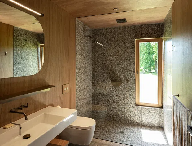 A compact, refined bathroom with a walk-in shower clad in small speckled stone-like tiles, a wall-mounted toilet, and a sculptural white basin on timber joinery. Warm timber panels cover the ceiling and walls, complemented by a rounded wall mirror, black tapware, a timber-framed window, and a heated towel rail. Natural light and greenery are visible through the window.