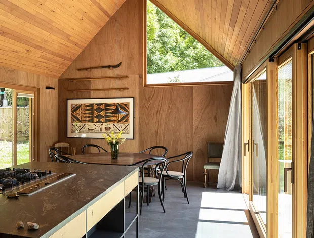 A warm, timber-lined open-plan kitchen and dining space with a dramatic sloping, vaulted ceiling. The room features a dark stone island with open shelving, a dining table and chairs, extensive full-height glazed doors, a triangular clerestory window, polished concrete flooring, and a sculptural ceiling fan. Strong sunlight enters through the glazing, creating a bright connection to the outdoors.
