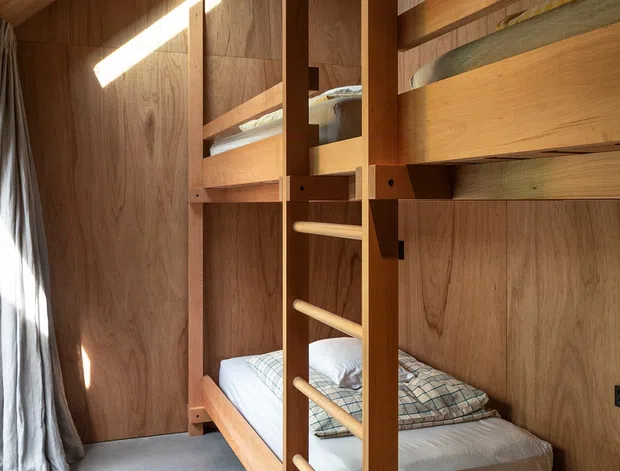 A compact bunk bedroom lined throughout with warm timber wall and ceiling panels. Built-in wooden bunk beds with integrated ladder occupy the right side, with neatly made beds, plaid bedding, and a small high-level skylight casting strong geometric sunlight across the timber surfaces. A grey polished floor and full-length curtain complete the simple, warm sleeping space.