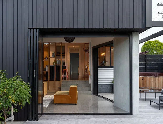 Contemporary residential exterior with a large open glazed entry and dark vertical cladding. The view looks into a warm, refined interior featuring timber shelving, polished concrete or stone flooring, and a mustard lounge chair. A covered outdoor terrace with seating and timber screening is visible to the right, alongside a concrete structural wall and overhead beams.
