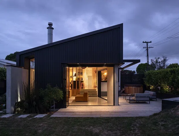 A compact contemporary dark-clad residential extension or backyard dwelling photographed at dusk. The angular single-storey structure has a steep asymmetrical sloping roof, black vertical cladding, a chimney, and extensive glazed openings revealing a warmly lit interior. A timber deck forms an outdoor entertaining area with lounge seating and a dark rectangular outdoor feature in the garden.