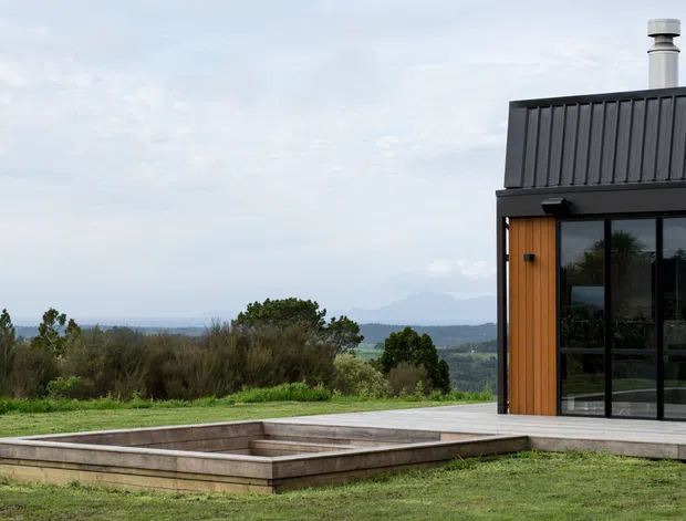 Contemporary dark-clad house exterior set on a grassy elevated site, with a low rectangular timber-edged sunken feature or pool in the foreground. The building has a steeply pitched standing-seam metal roof, black-framed floor-to-ceiling glazing, and a warm timber-clad entry door. Rolling rural or coastal landscape is visible beyond.