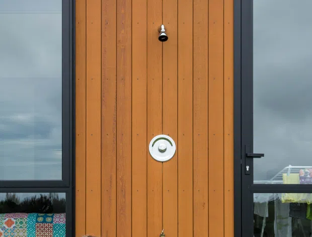 Close-up exterior view of a contemporary house entrance, featuring a tall warm timber-clad central door or wall panel framed by dark charcoal aluminium glazing and doors. A child sits on the pale outdoor deck beside the entrance. The facade has clean horizontal and vertical lines, integrated door hardware, and large glazed openings reflecting the overcast sky and surrounding yard.